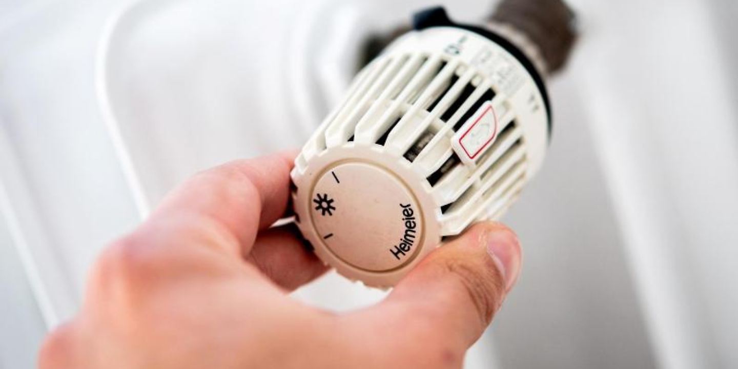 Ein Mann dreht in einer Wohnung am Thermostat einer Heizung. 
